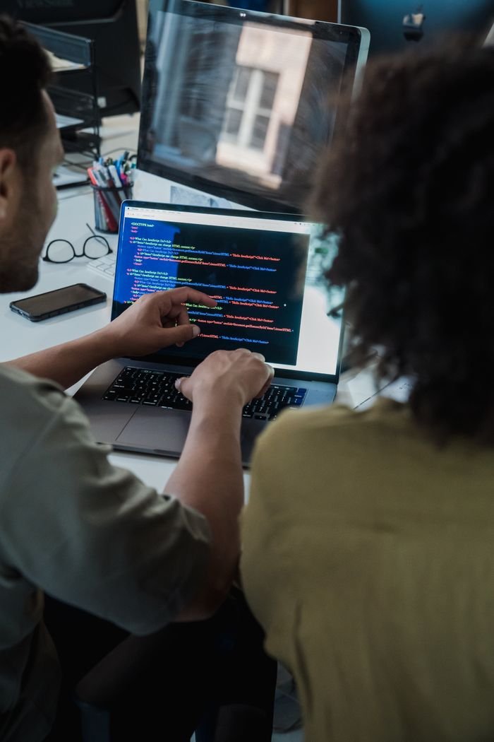 Contact Two adults collaborating on programming tasks using a laptop in an office setting.