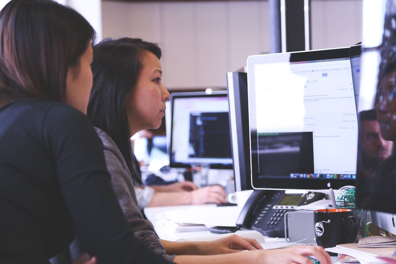 Home Two women working together on code at a computer in a modern office setting.